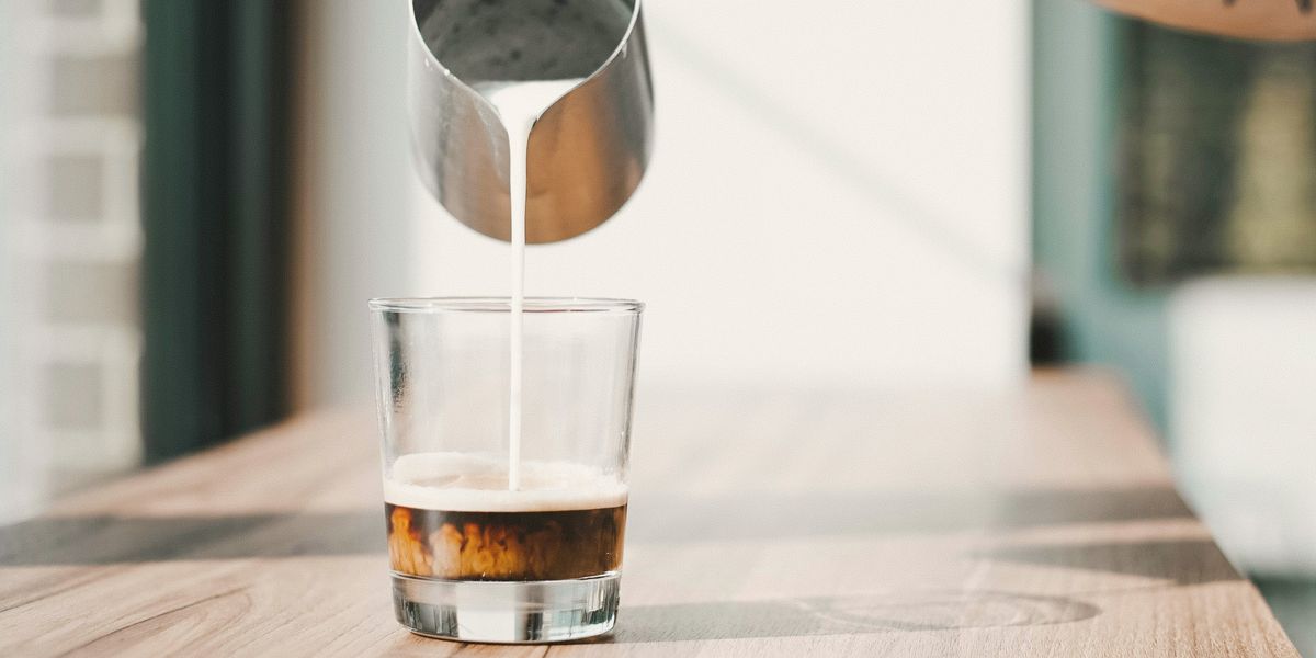 A barista's tattooed arm pouring steamed milk into a short glass of espresso on a timber bench by the window.