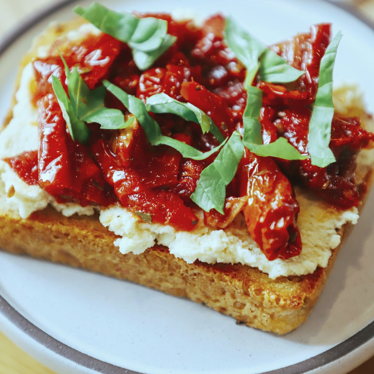 A thick slice of toast spread with whipped feta and piled with roasted tomatoes and torn basil, on a grey-rimmed plate on a timber table.