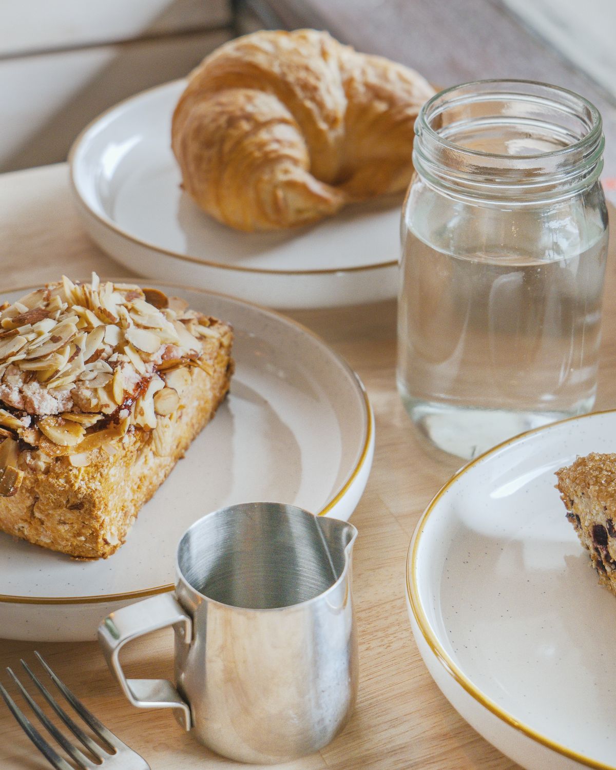 An almond croissant and a plain croissant on white plates, with a small milk jug and a glass of water on a timber table.