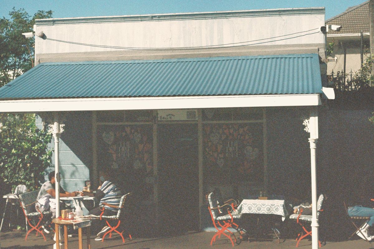 A single-storey café under a blue corrugated-iron verandah on white posts, with people at small tables on the footpath in the morning sun.