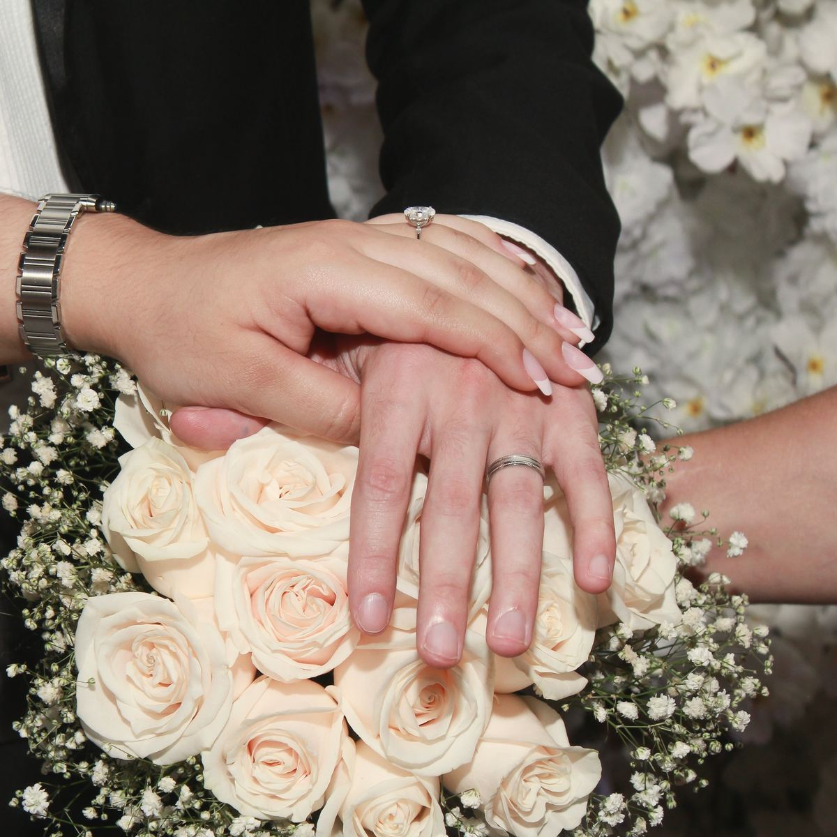 Hands laid one over another on a bouquet of pale roses, two wedding rings showing.