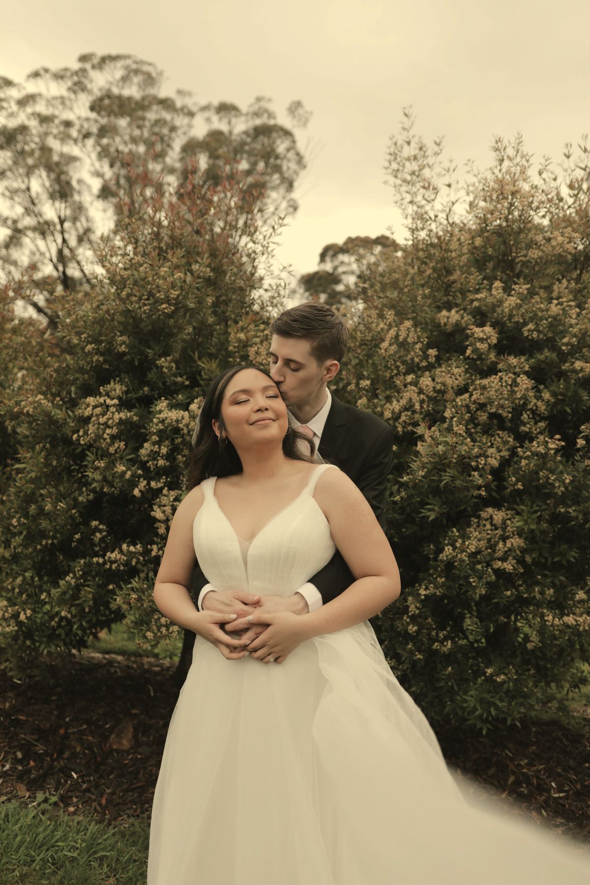 Callum holding Priya from behind among the flowering shrubs in the last warm light.