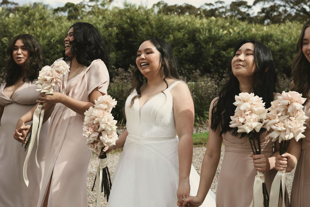 Priya laughing with her bridesmaids in dusty pink, all holding pale bouquets.