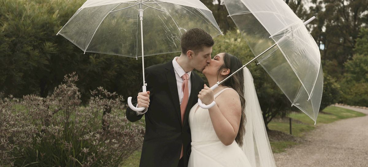 Priya and Callum kissing under two clear umbrellas on the garden path.