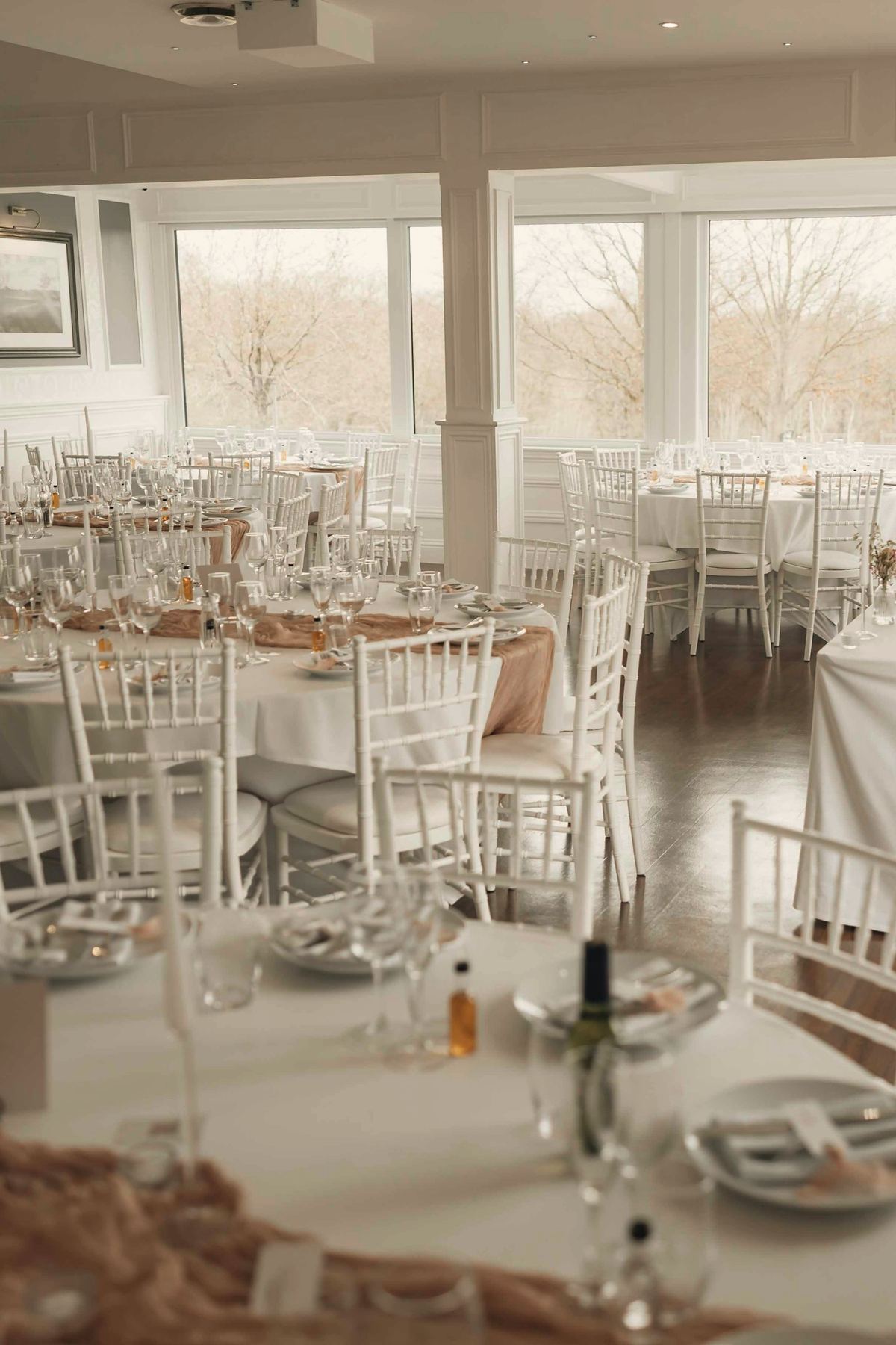 The hall set for lunch, round tables and white chairs by tall windows onto the trees.