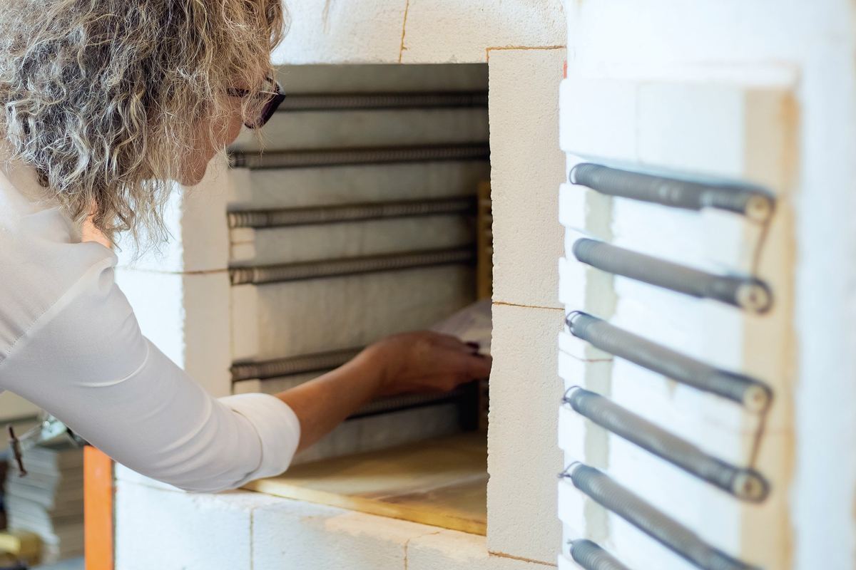 A potter reaching into the open kiln to set a piece on the shelf, its elements lining the door.