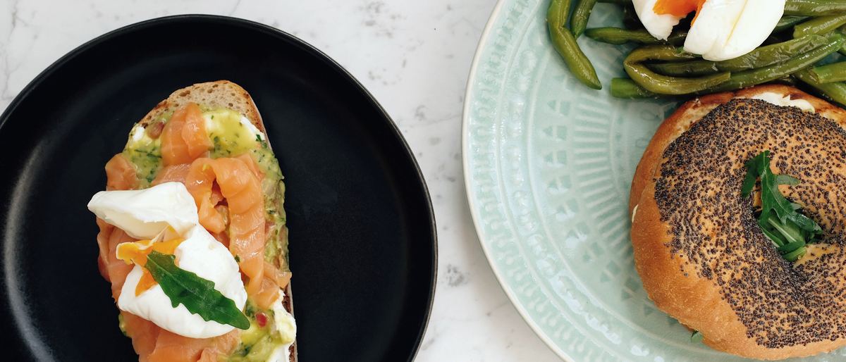 Brunch plated on handmade stoneware: salmon toast with a poached egg on a near-black plate, a bagel with green beans on a carved celadon plate.