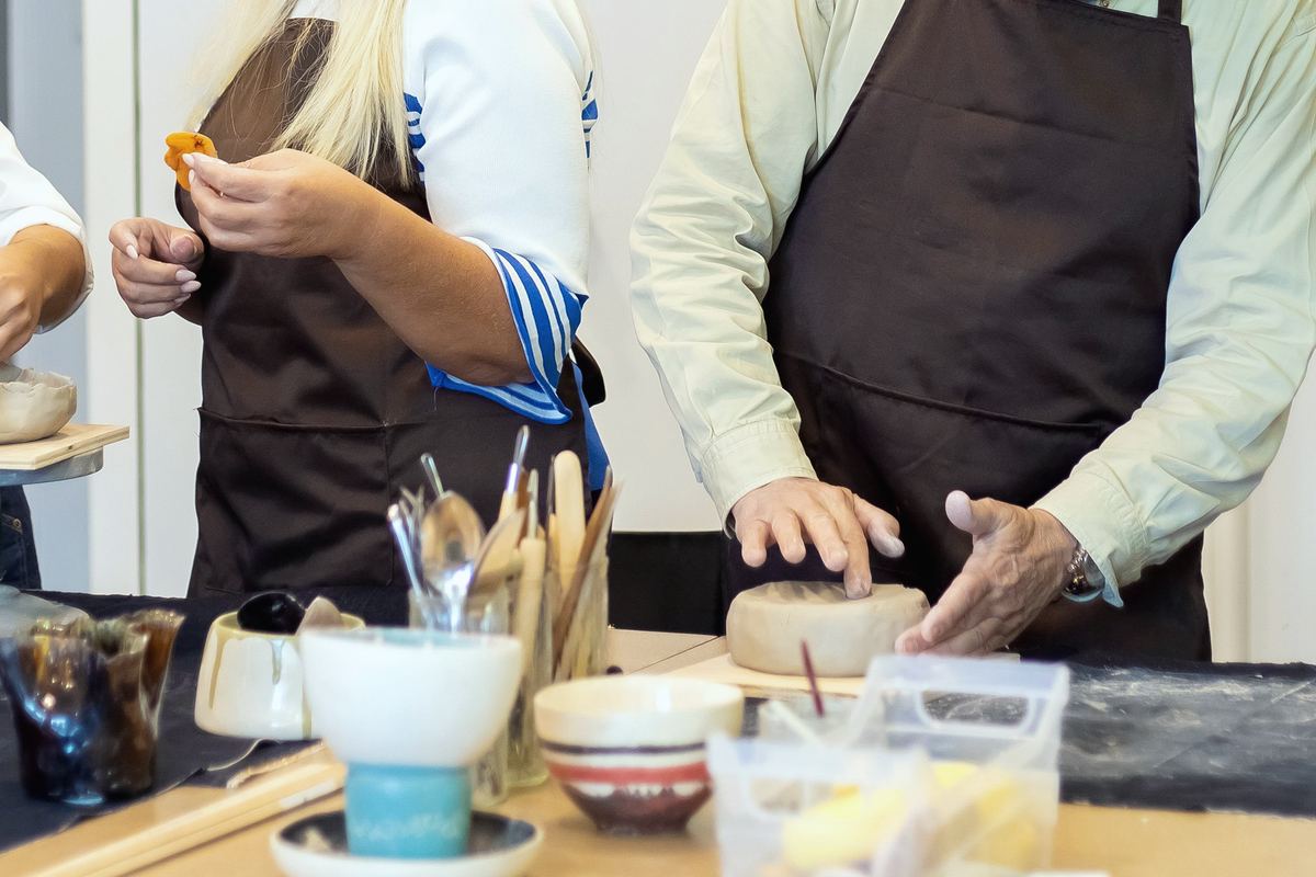 Hands at the big bench: two people in aprons shaping small pots, with tools, water and glaze pots between them.