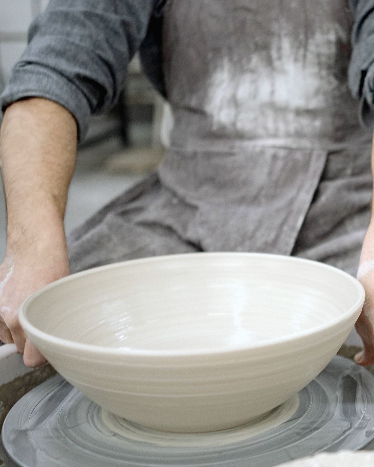 Hands steadying a wide, freshly thrown bowl on the wheel, the throwing lines still wet.