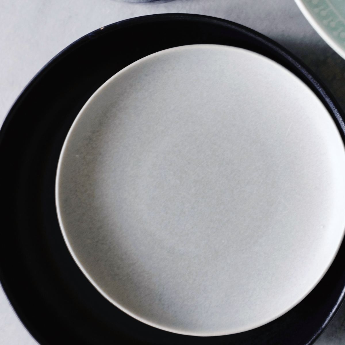 A matte off-white dinner plate from above, its whole rim showing against a black plate.
