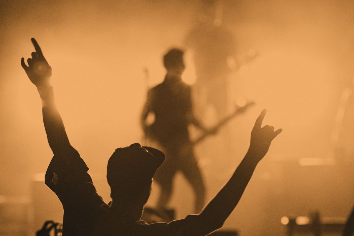 A crowd from behind with arms up, facing a guitarist on a small, smoky stage.