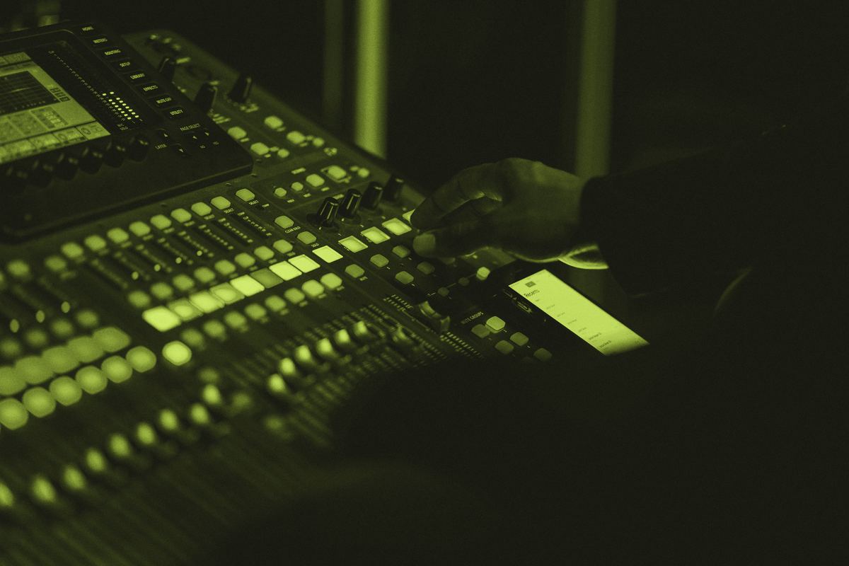 A hand on the faders of a lit mixing desk in a dark room.