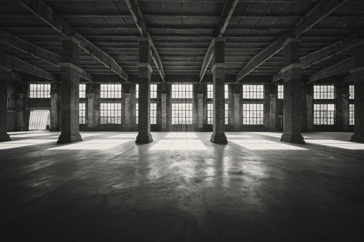 An empty old warehouse floor: brick columns, timber beams and a row of tall windows letting sunlight in.