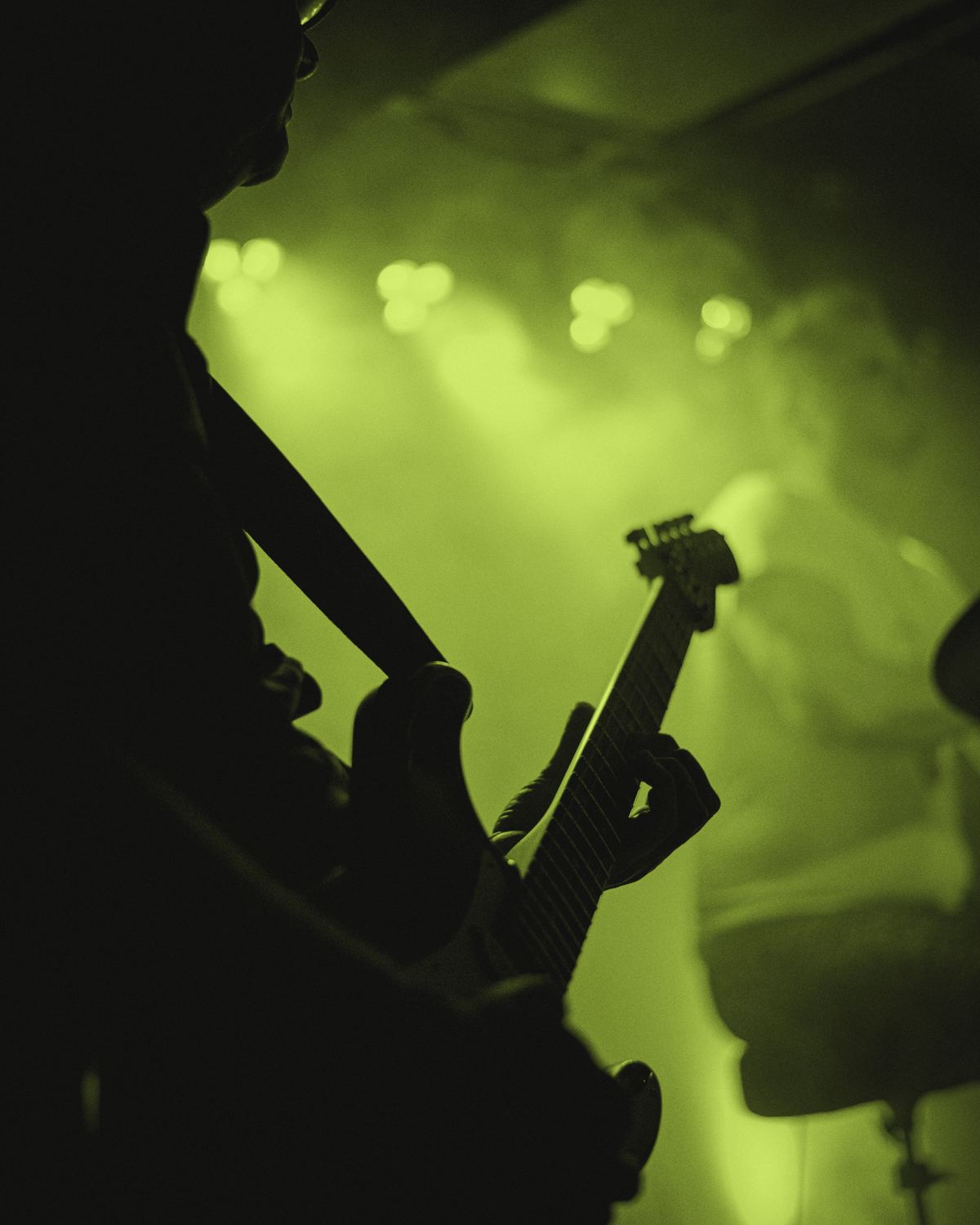 A guitarist seen from behind in silhouette, playing into stage lights and haze.