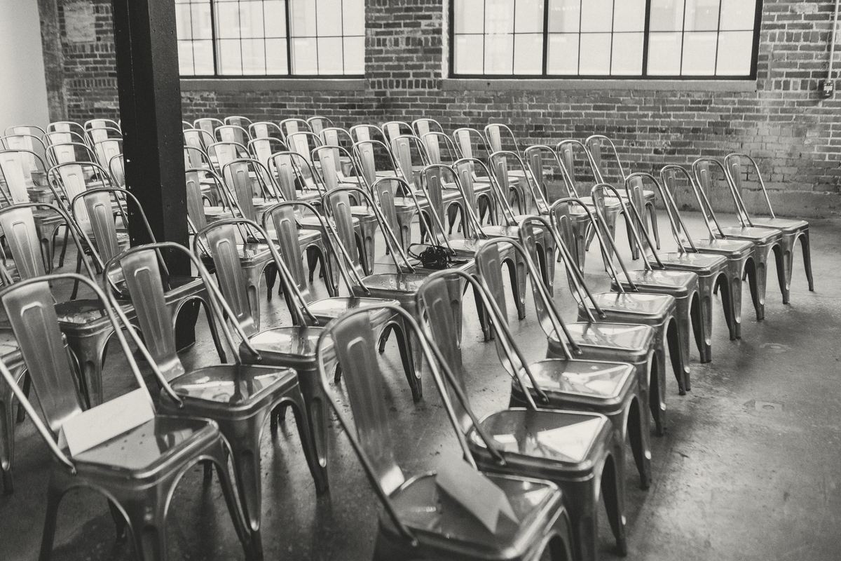 Rows of metal chairs set out in a brick-walled warehouse room with tall steel windows.