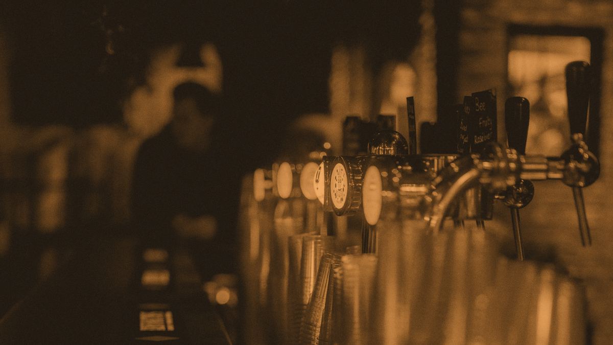 A row of beer taps along a dark bar, with stacked cups behind the counter.